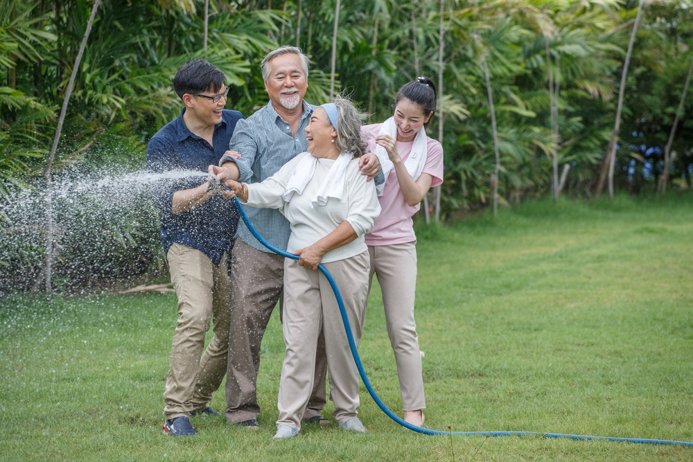 An elderly woman watering a garden with a hose. She is laughing and is surrounded by an elderly man, and a younger man and woman, all smiling.